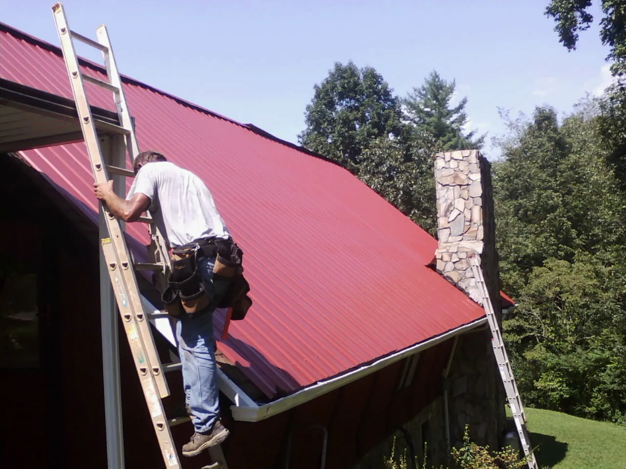 Roofer on a ladder beside a red metal roof and stone chimney — Soffit & Fascia Repair by Roofing Brothers in Chaptico